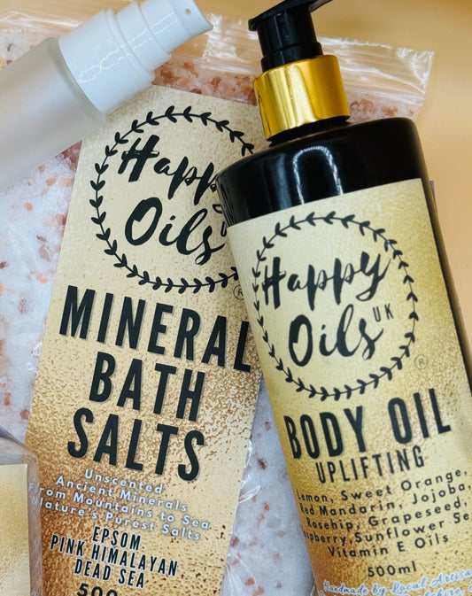 Happy Oils UK bath and body products including a 500g bag of mineral bath salts and 500ml body oil uplifting and 30ml travel bottle on a yellow background.