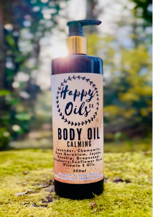 A 500ml bottle of Happy Oils Body Oil Calming, featuring a black and gold pump dispenser, set against a blurred forest background on green moss.