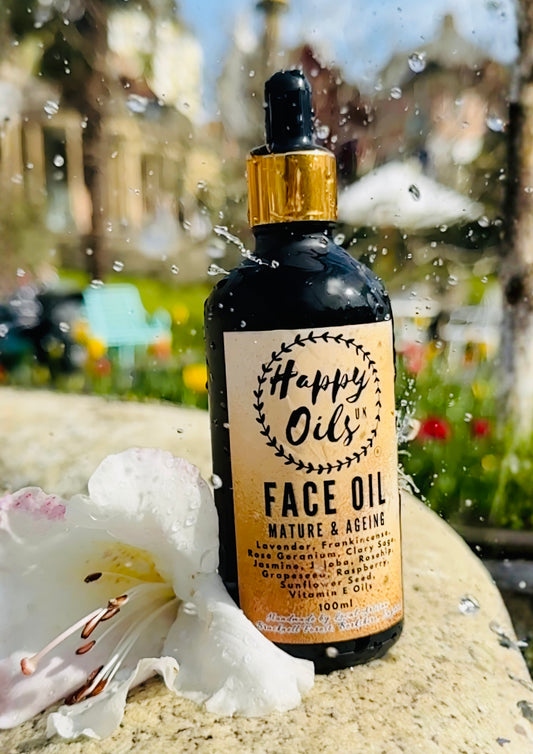 A bottle of Happy Oils UK Face Oil with a gold cap and beige label, placed outdoors on a stone surface with a white flower in the foreground and water droplets splashing around it.