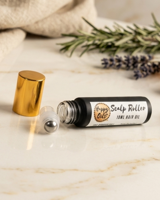 A small roll-on hair oil bottle labeled 'Happy Oils UK Scalp Roller" lies open on a light marble surface, with its gold cap removed and placed beside it. Rosemary sprigs and a beige cloth are softly blurred in the background.