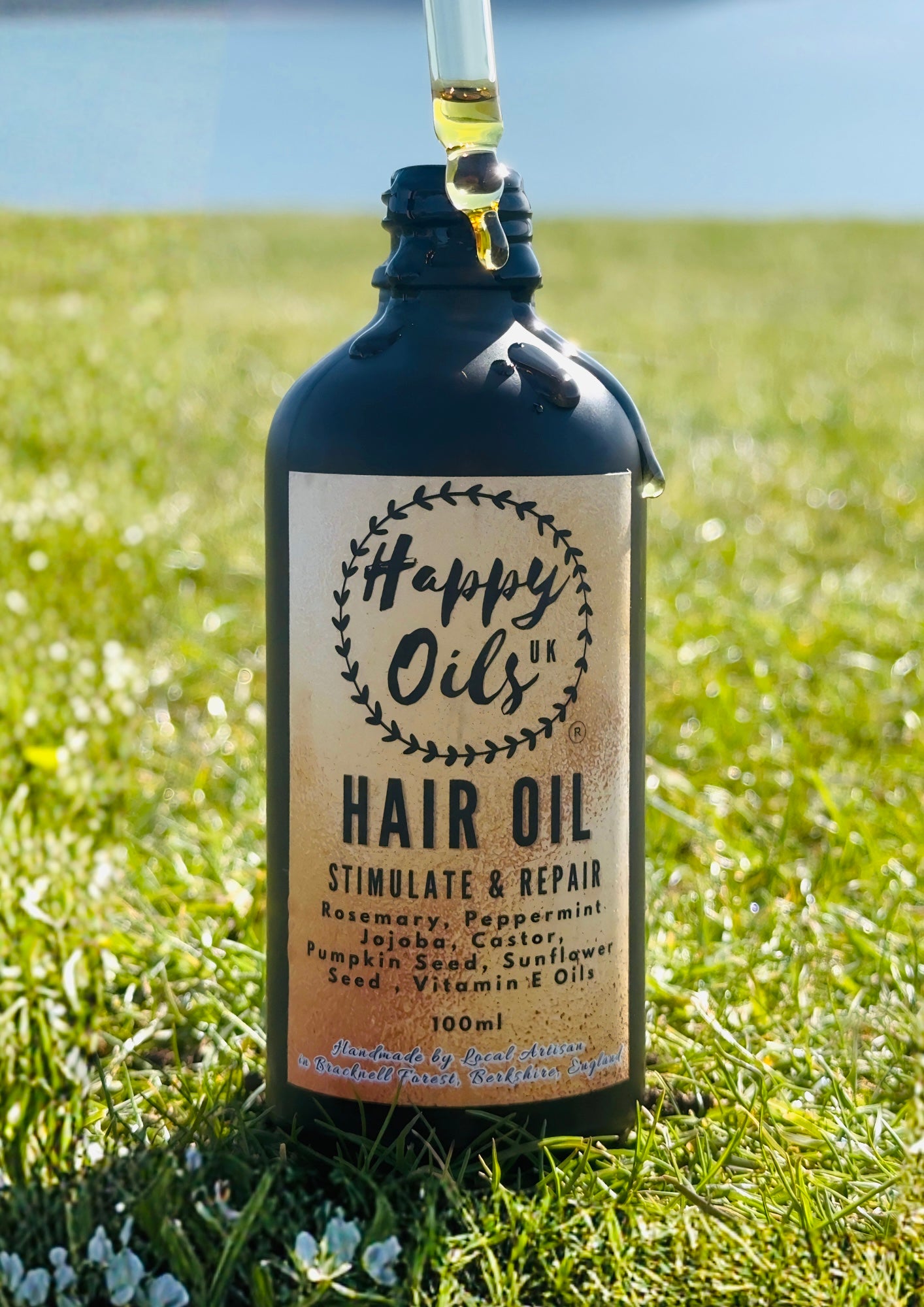 A bottle of Happy Oils Hair Oil standing upright in a grassy outdoor field, with oil being poured into the open bottle. The label reads 'Happy Oils Hair Oil' and lists ingredients such as rosemary, peppermint, jojoba, castor, pumpkin seed, sunflower, and vitamin E oils.