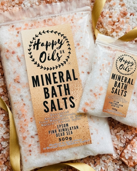 Pack of  Happy Oils UK Mineral Bath Salts, Unscented, Ancient Minerals, Pink Himalayan, Dead Sea, Epsom Salts 500g, arranged on a bed of coarse pink himalayan salt crystals with gold ribbons. The main 500h bag is in the foreground with the smaller 100g bag behind it.