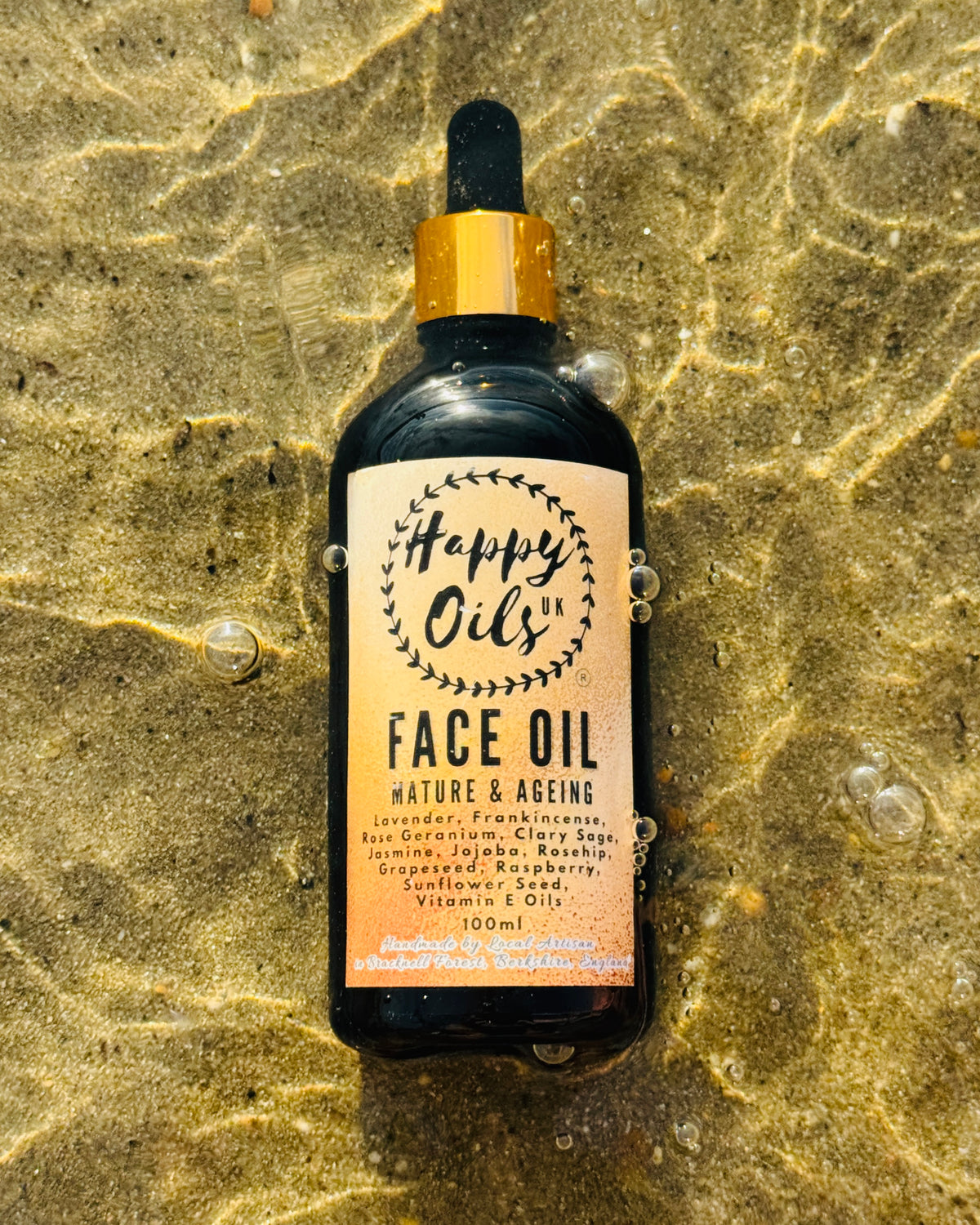 A 100ml bottle of Happy Oils UK Face Oil with a gold collar and black dropper cap, resting on wet, sunlit sand with water droplets around it.

Ingredients on label: lavender, Frankincense, rose geranium, clary sage, jasmine, jojoba, rosehip, red raspberry, grape seed, sunflower seed, vitamin E Oils

Handmade in Bracknell Forest, Berkshire, England