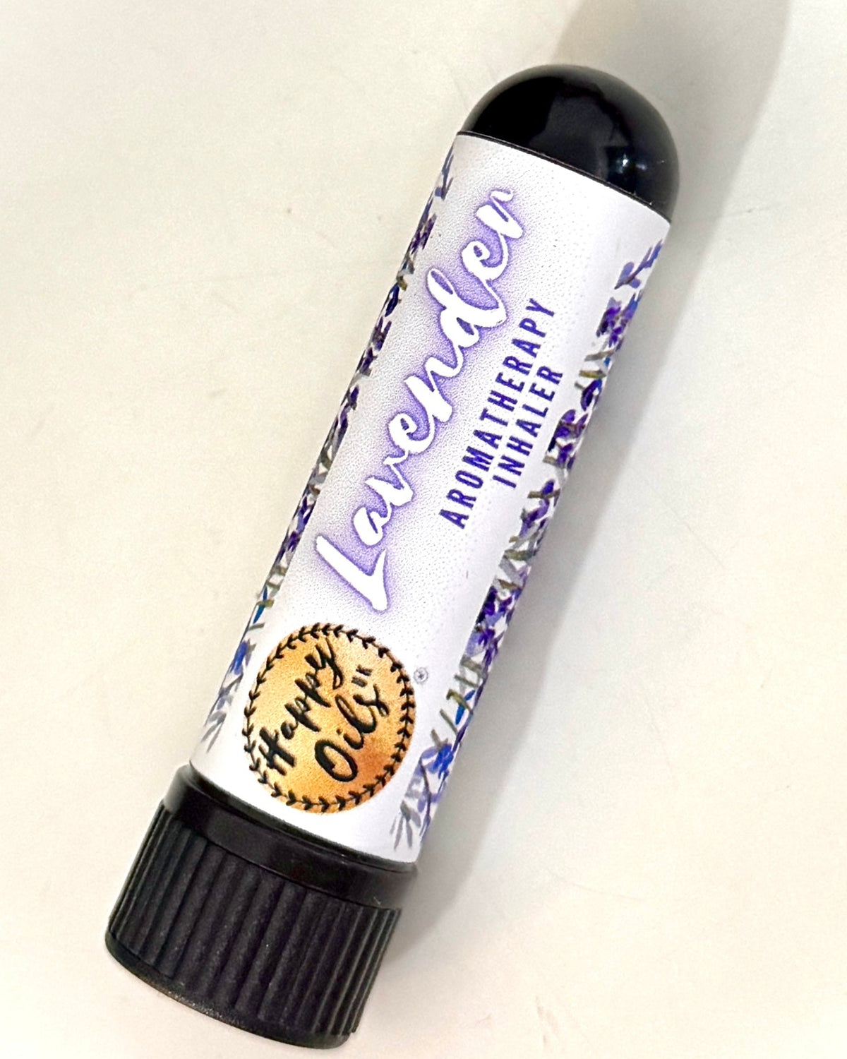 A tube of Happy Oils UK Lavender Aromatherapy Nasal Inhaler on a plain off-white background, angled diagonally. The tube has a white label with purple lavender illustrations, purple 'Lavender' text, and a gold circular seal, topped with a black cap.