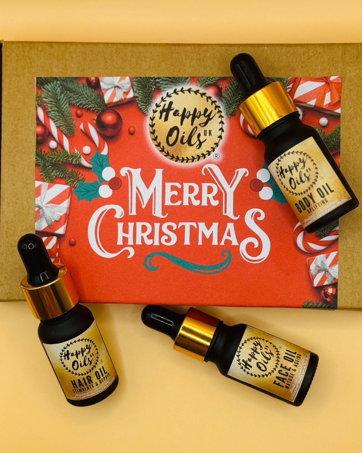 Three 10ml mini bottles of 'Happy Oils UK' hair oil face oil and body oil with a decorative Christmas box in the background.