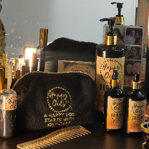 Set of hair and skin care products including hair oil, face oil and body oil bottles, a comb, and a black pouch with gold emroidered logo of Happy Oils UK and text saying A happy you starts with self care on a wooden surface with a blurred background.