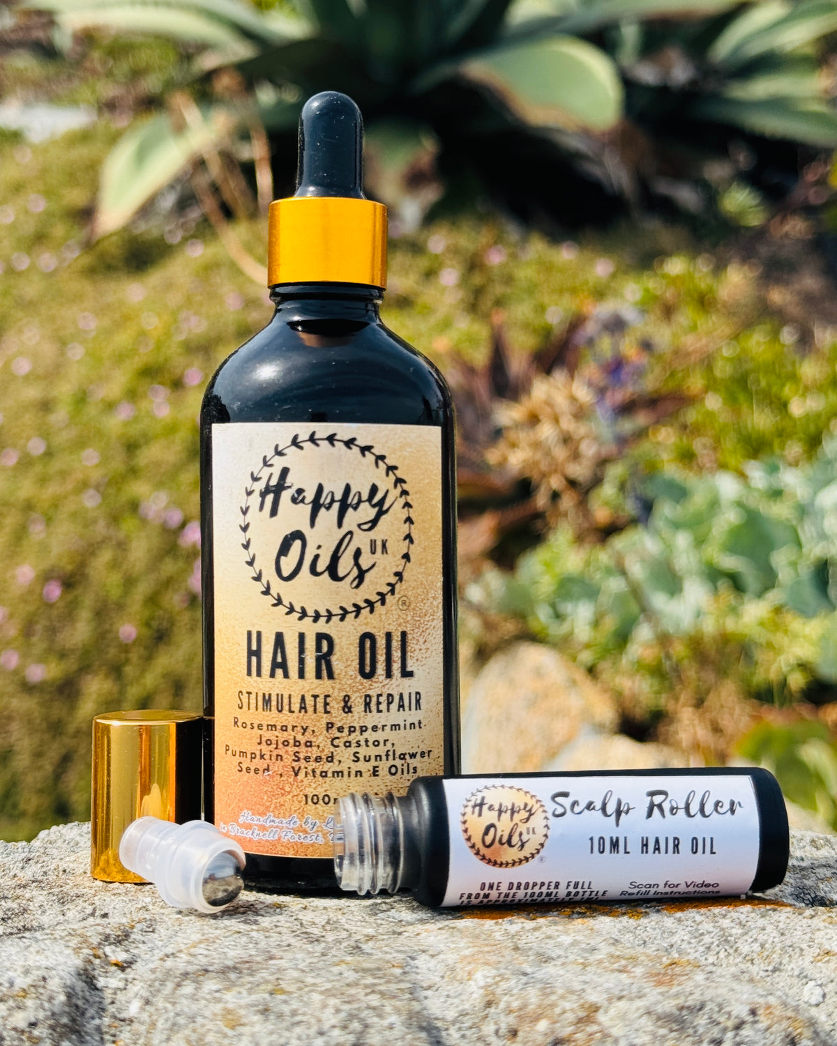A bottle of 100ml Happy Oils UK Hair Oil stands upright on a stone surface outdoors, with a gold dropper cap and a separate opened scalp roller bottle lying beside it. The background shows blurred greenery and plants.